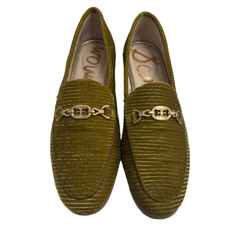 A pair of olive green ribbed velvet loafers with gold-tone chain link detailing across the vamp, displayed on a white tufted surface against a dusty pink background.