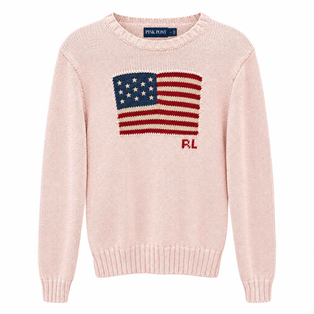 A pale pink knit sweater hanging on a black hanger, featuring a knitted American flag graphic on the chest with red and blue details and the letters 'RL' below. The background shows a clothing store interior with other garments and a wooden floor.